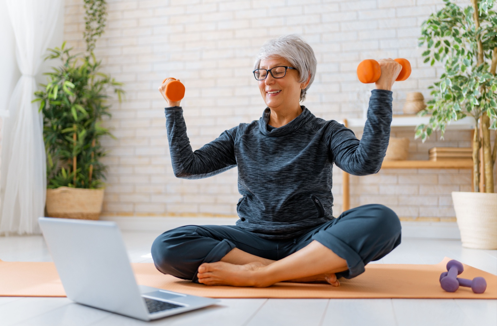 A happy senior wears athletic clothing follows along with an exercise video with their dumb bells.
