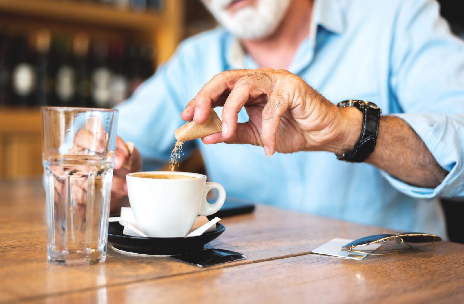 A close-up image of an older adult in a coffee shop pouring artificial sweetener into their coffee.