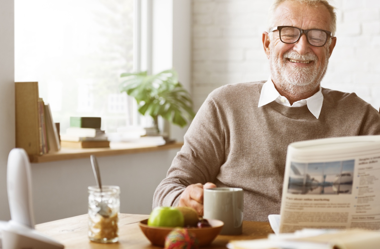 An older adult laughing while drinking coffee and reading their newspaper in the morning with a bowl of fruit in front of them.
