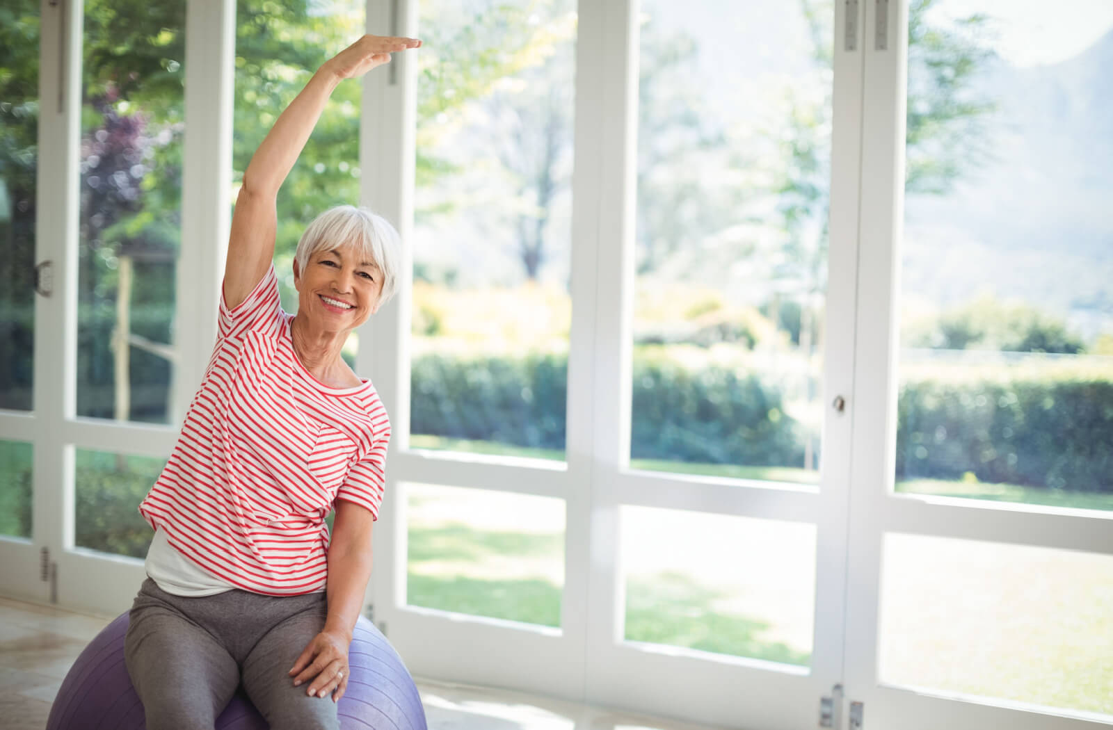 An older adult stretching an arm overhead in front of a sunlit window while cooling down after a core exercise class.
