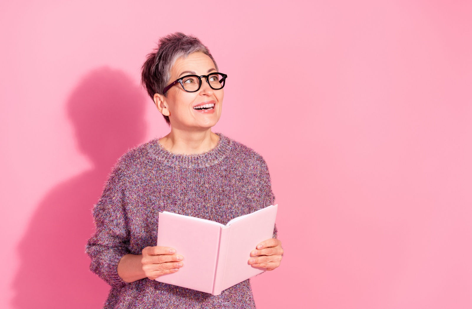 Against a pink backdrop, a senior in a wool sweater holds a book, looking up in excitement over how it’s making them think