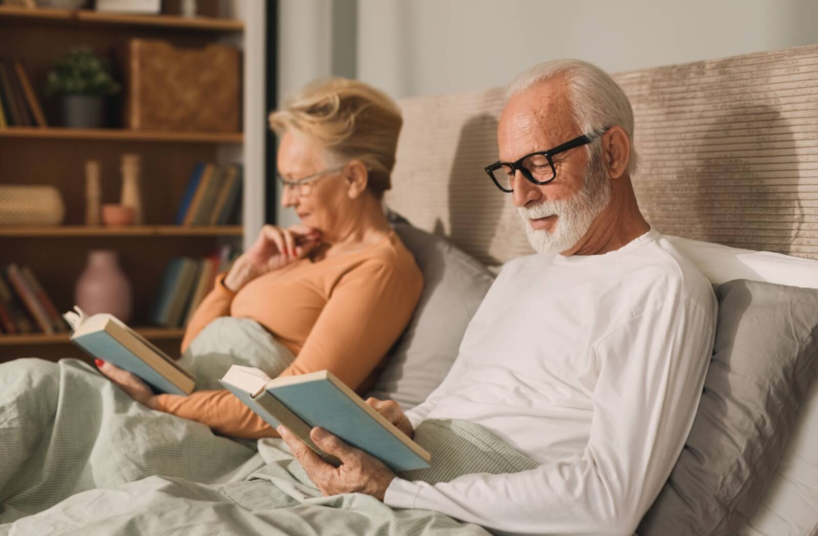 A senior couple sit beside each other in bed, reading two copies of the same book as part of their bedtime routine