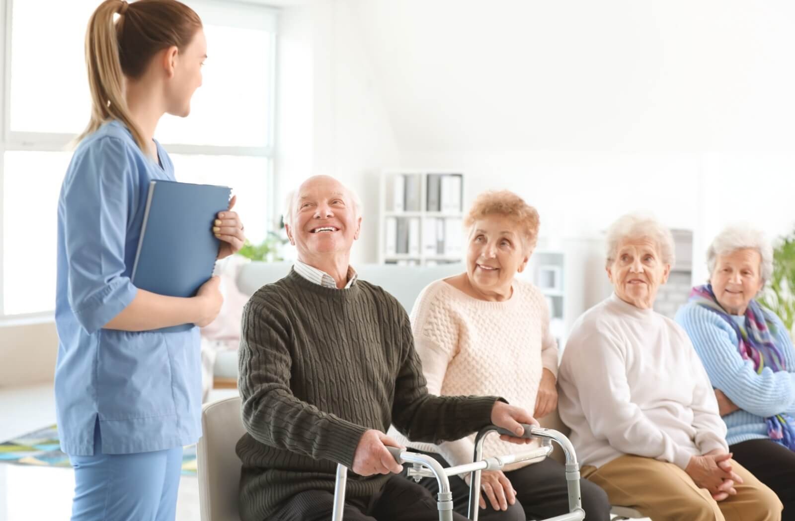 An older adult sits beside their friends in respite care and smiles up at a caregiver during a regular check
