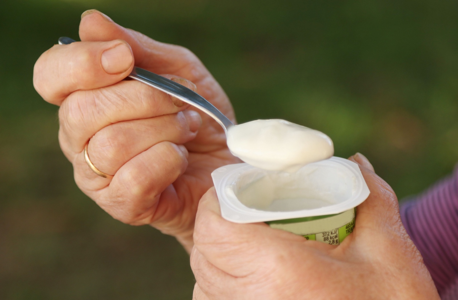 A close-up view of an older adult holding a yogurt cup in their left hand and a spoonful of yogurt in their right hand.