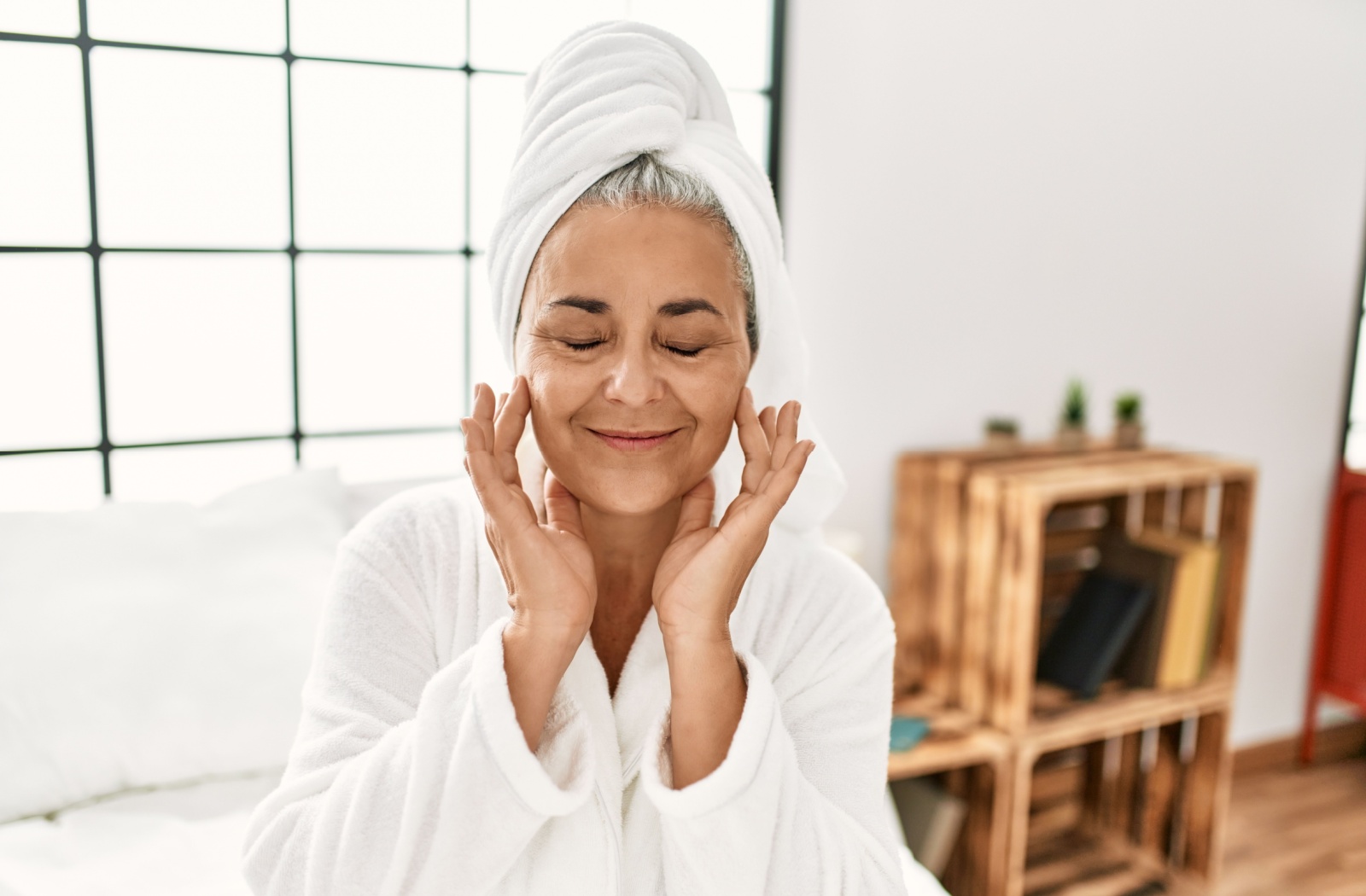 An older lady in a bath robe applies moisturizer to her face after her shower.