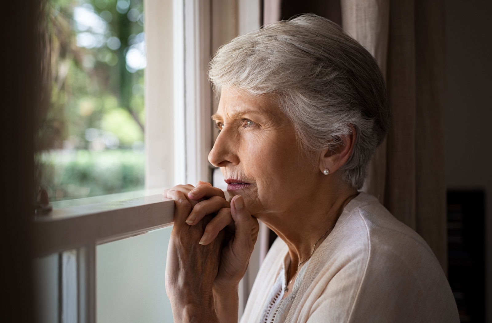 An older adult woman holding onto a window sill and looking out the window with a serious expression.