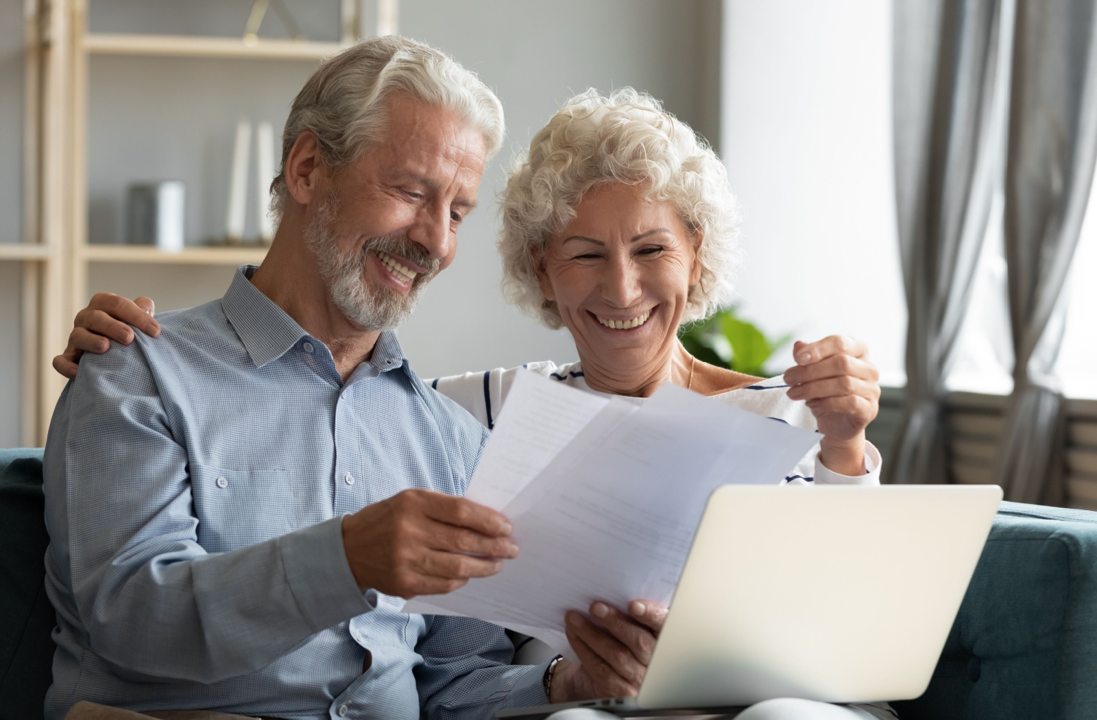 A smiling older couple holding up paperwork in front of an open laptop, happy they meet the physical requirements for a move to assisted living.