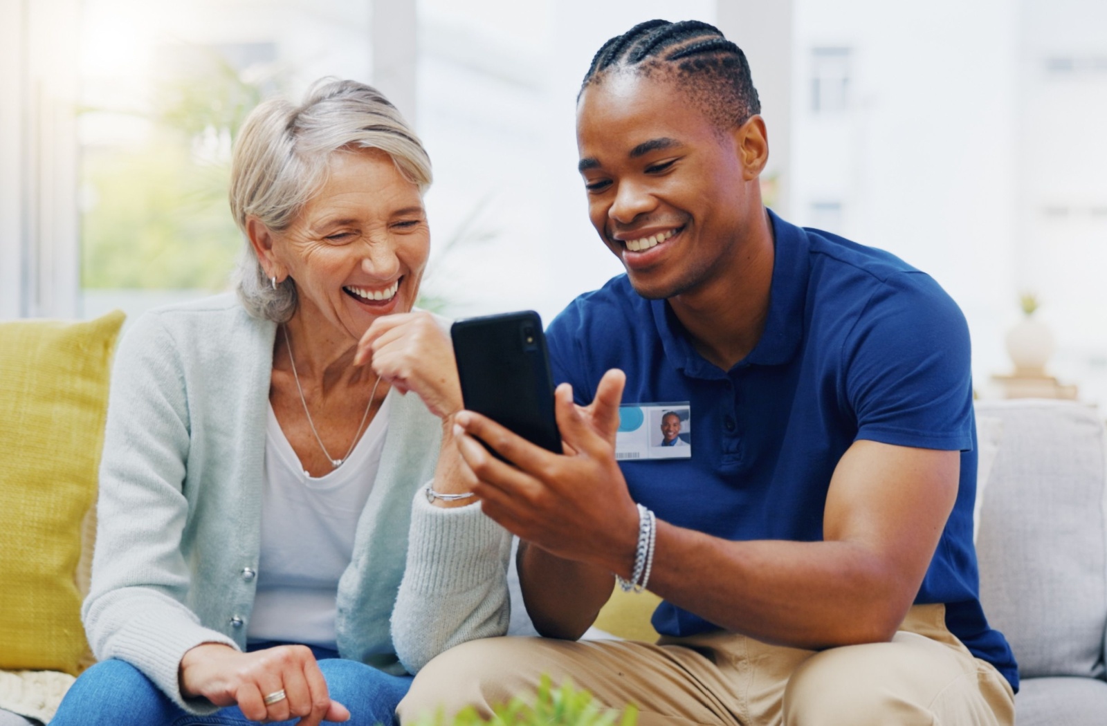 A caregiver sitting on a couch with an older adult and showing them something as they both smile at a smartphone.