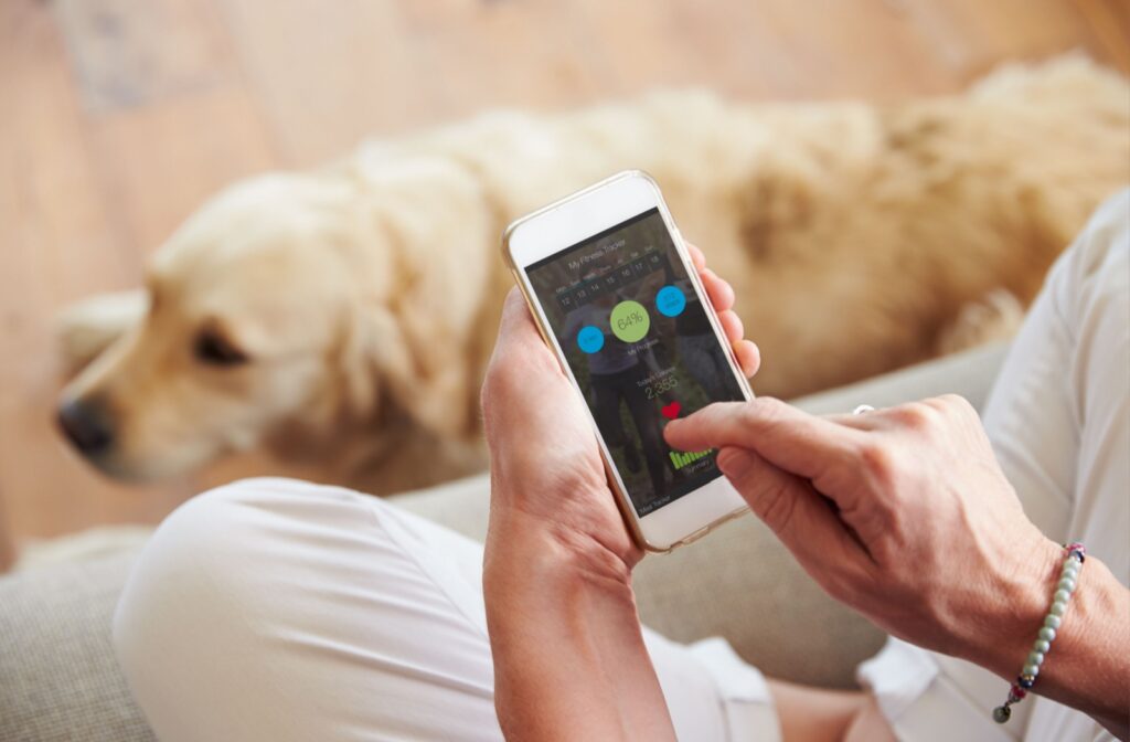 Close-up of a person checking a health-monitoring app on their phone while a dog rests on the floor beside them.