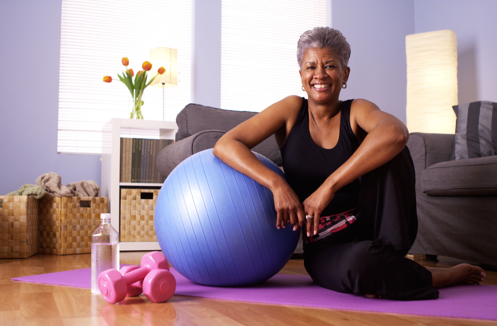 An smiling senior takes a picture with their workout equipment; a balance ball, dumb bells, and a yoga mat.