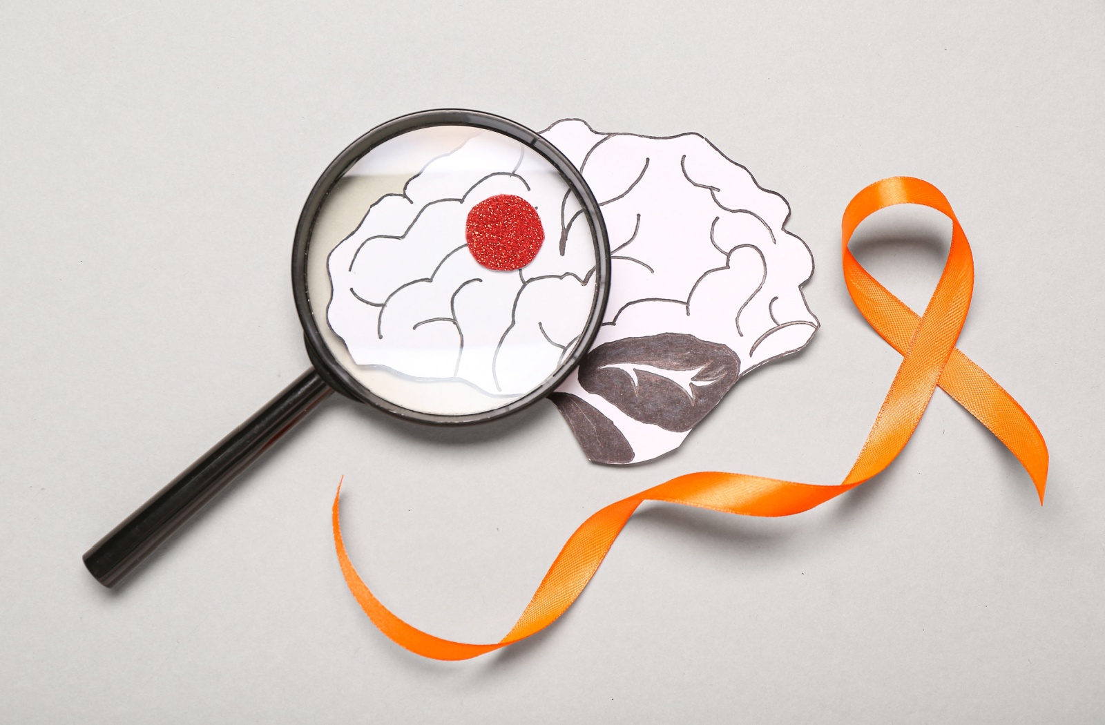 A magnifying glass rests over a drawing of a brain with a red dot, which, next to an orange ribbon, signifies MS